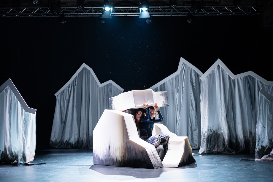 Bühnenbild erinnert an Eisschollen, aus den Eisschollen wird eine Art Höhle gebaut. Darin sitzen 3 Tänzer*innen.
