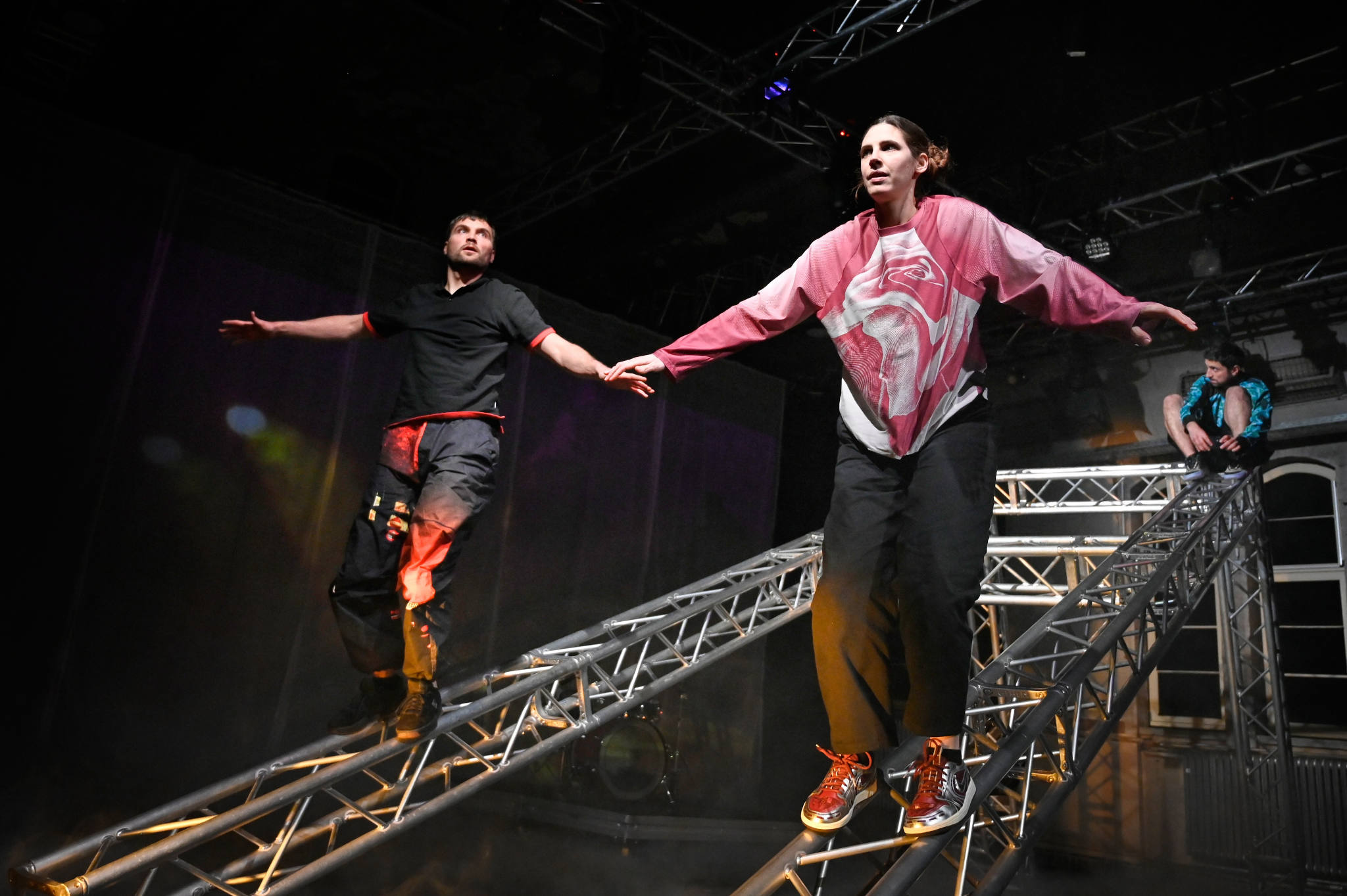 Zwei Personen balancieren auf einem Gerüst, eine Person sitzt auf dem selben Gerüst und filmt.